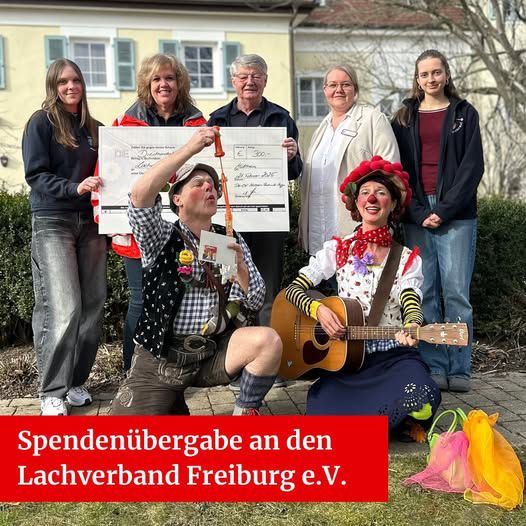 Spendenübergabe: Unterstützerinnen und Unterstützer mit Spendenscheck, davor zwei Clowns mit Gitarre